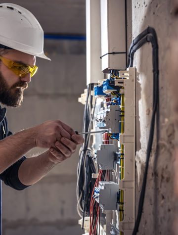 A male electrician works in a switchboard with an electrical connecting cable, connects the equipment with tools.